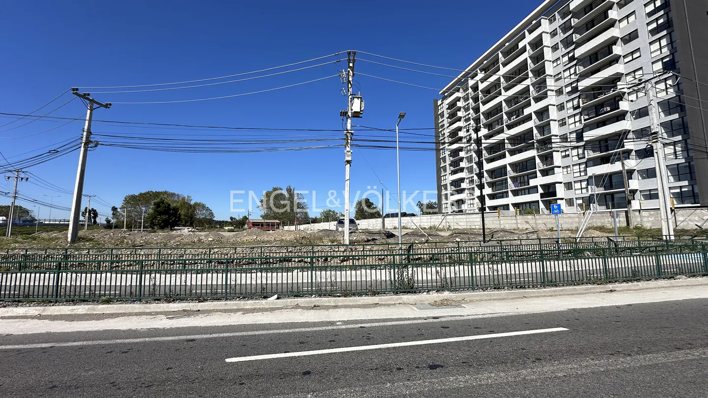 A vacant lot with a tall apartment building in the background under a blue sky. A road is in the foreground.