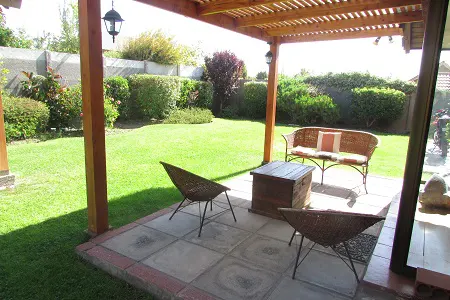 Outdoor patio with wooden pergola, wicker chairs, and a bench overlooking a green lawn and garden.