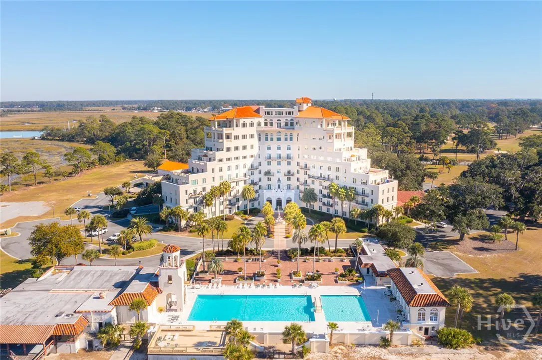 Aerial view of a white, multi-story resort with an orange tile roof, a pool, and palm trees.