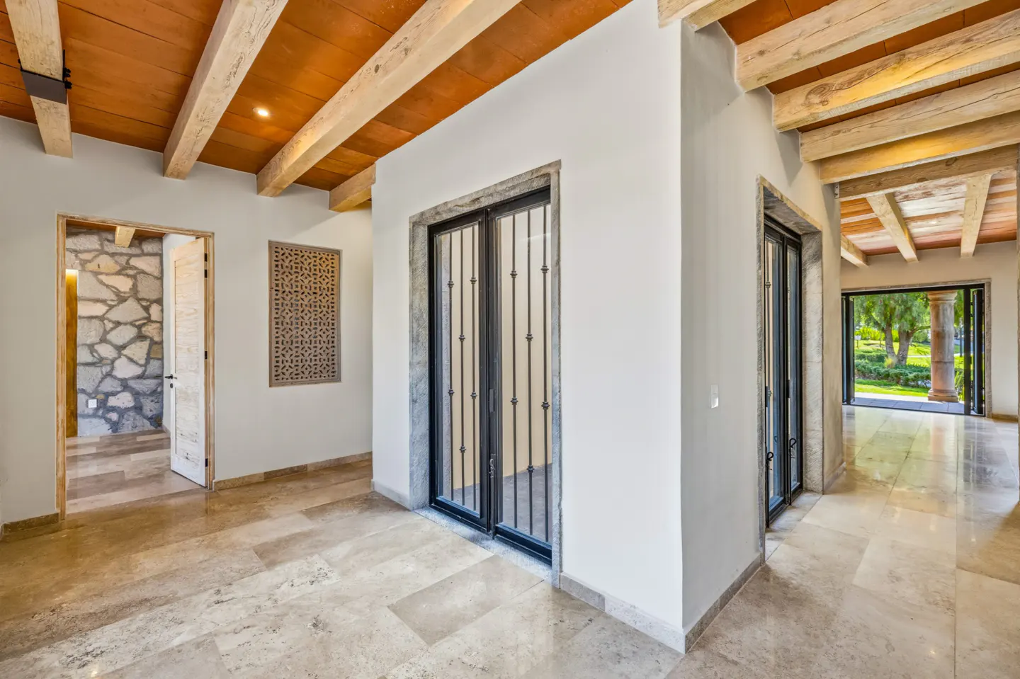 Hallway with travertine floors, white walls, and wood-beamed ceiling. Black metal doors lead to a garden view. A stone wall is visible through an open doorway.