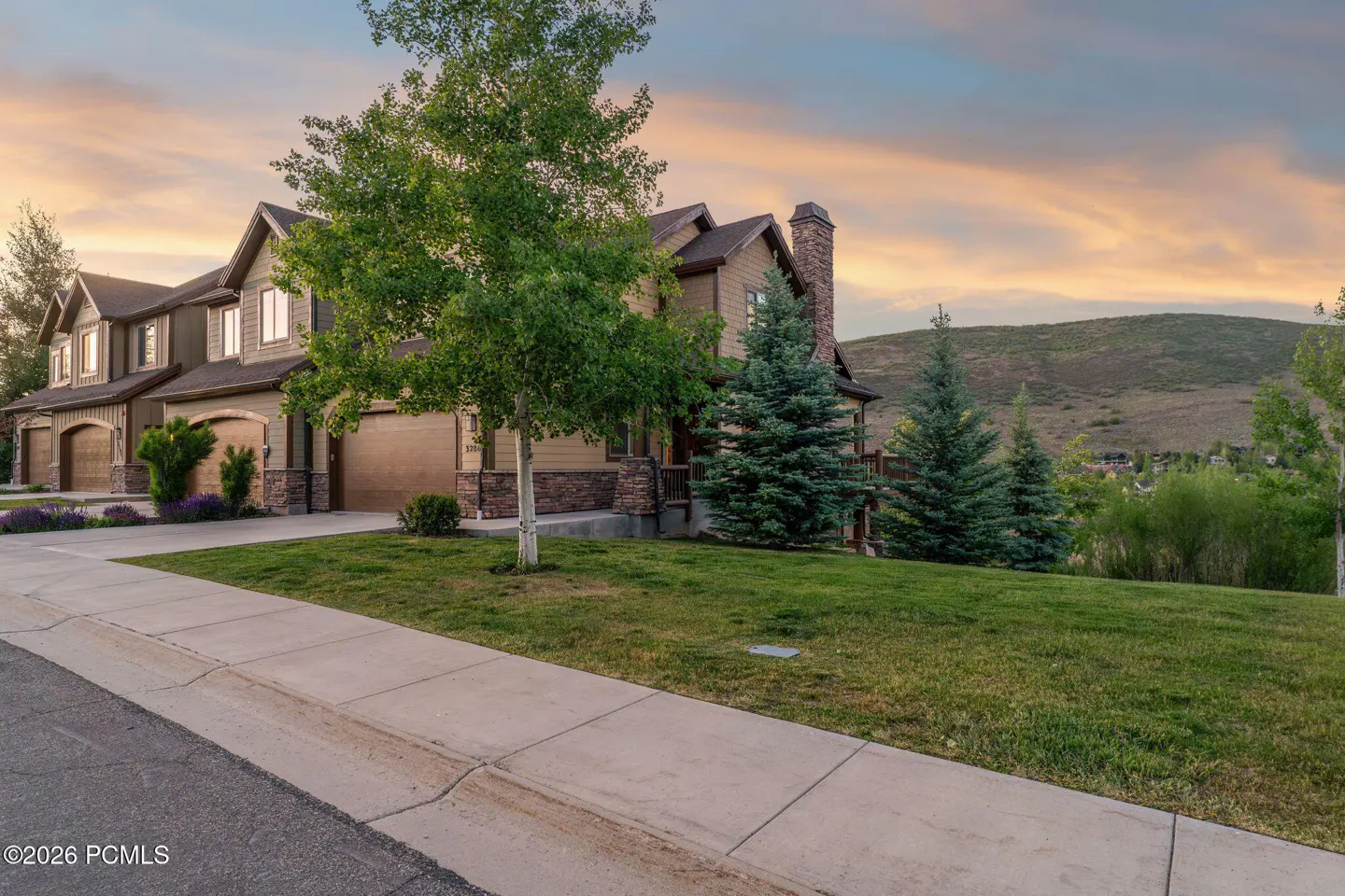 A multi-story tan home with stone accents and a garage at sunset, featuring a green lawn and mountain views.