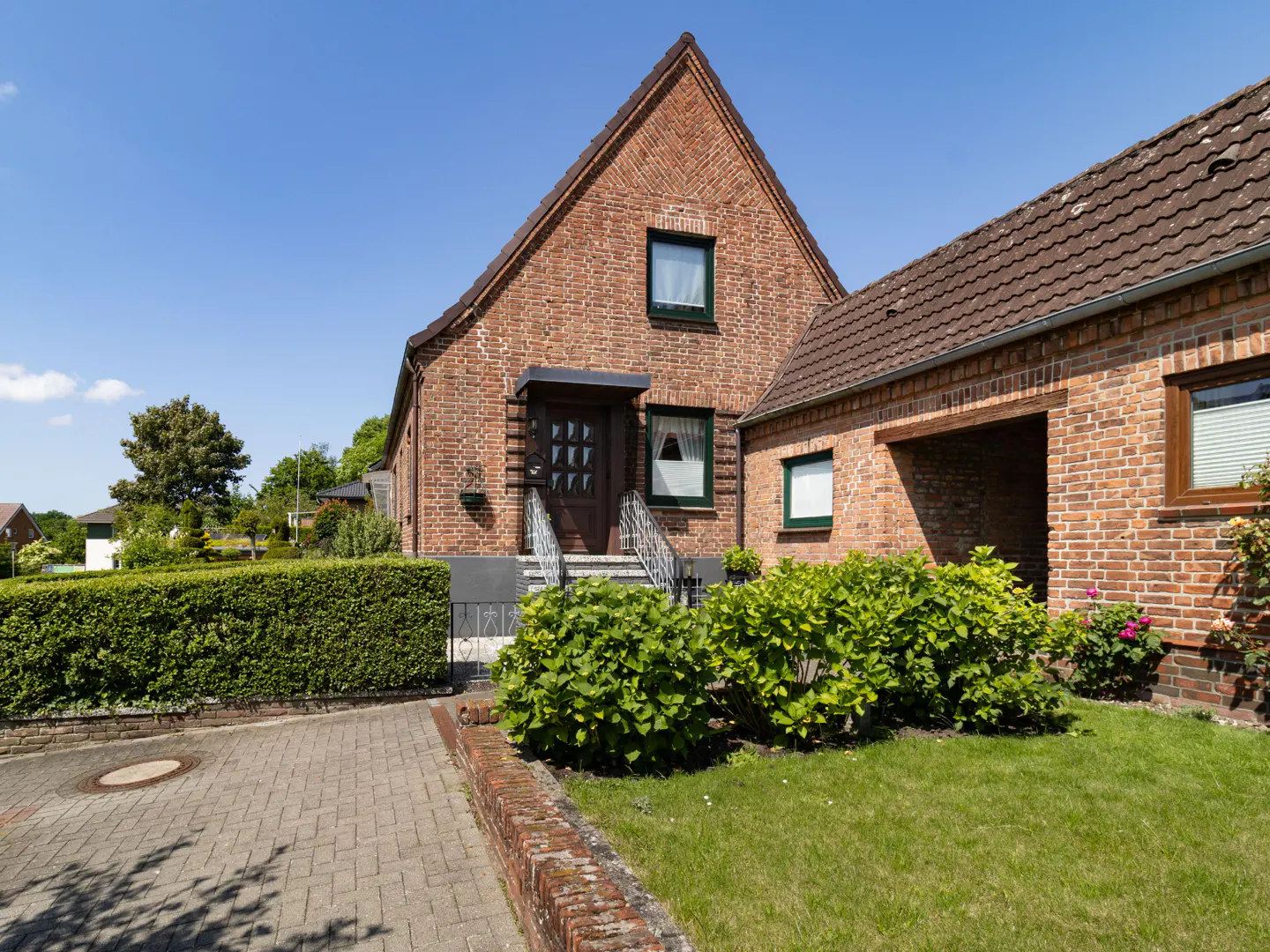 A two-story brick house with a gable roof, green trim, and a small front yard with green bushes.