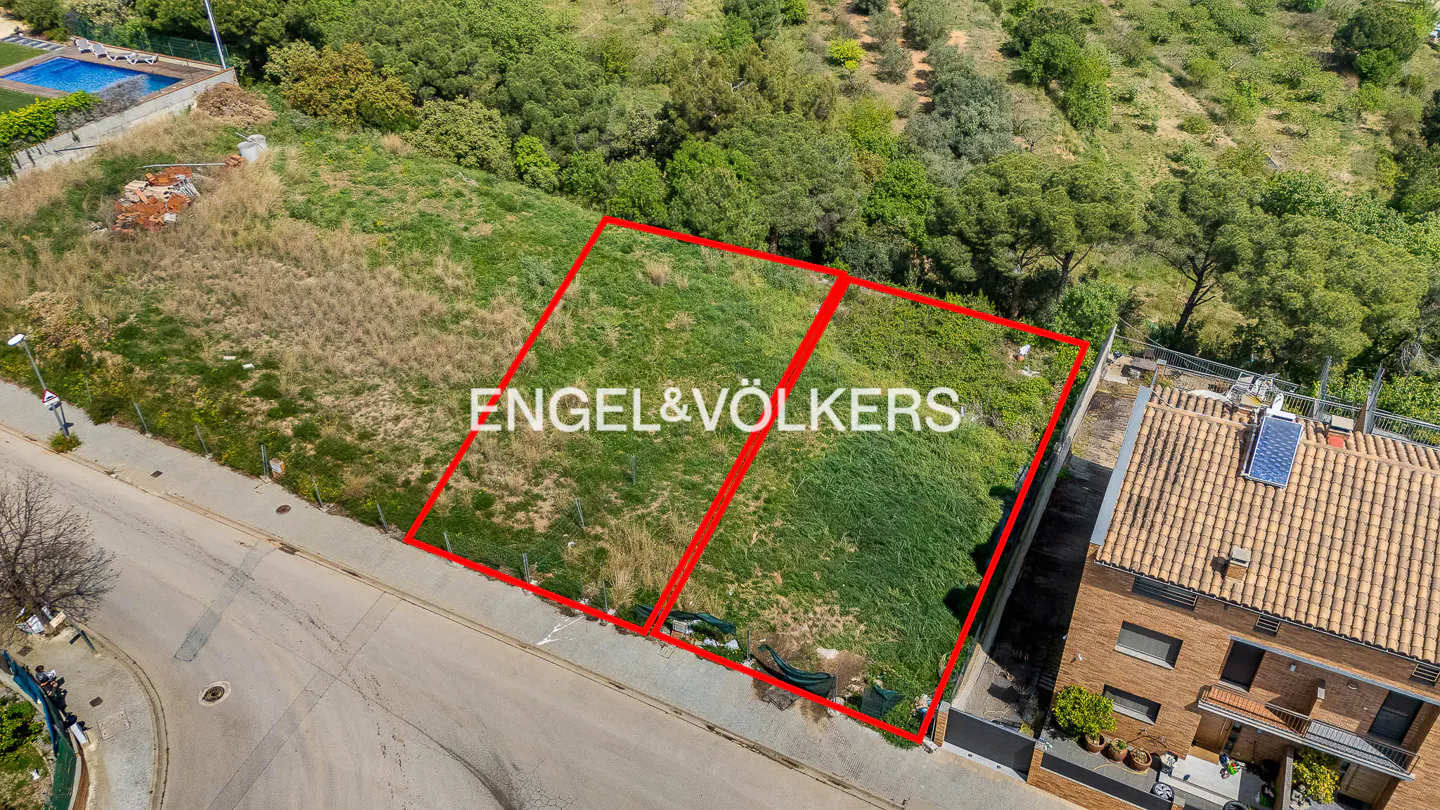 Aerial view of two adjacent grassy lots outlined in red, with Engel & Völkers logo, near a road and houses.
