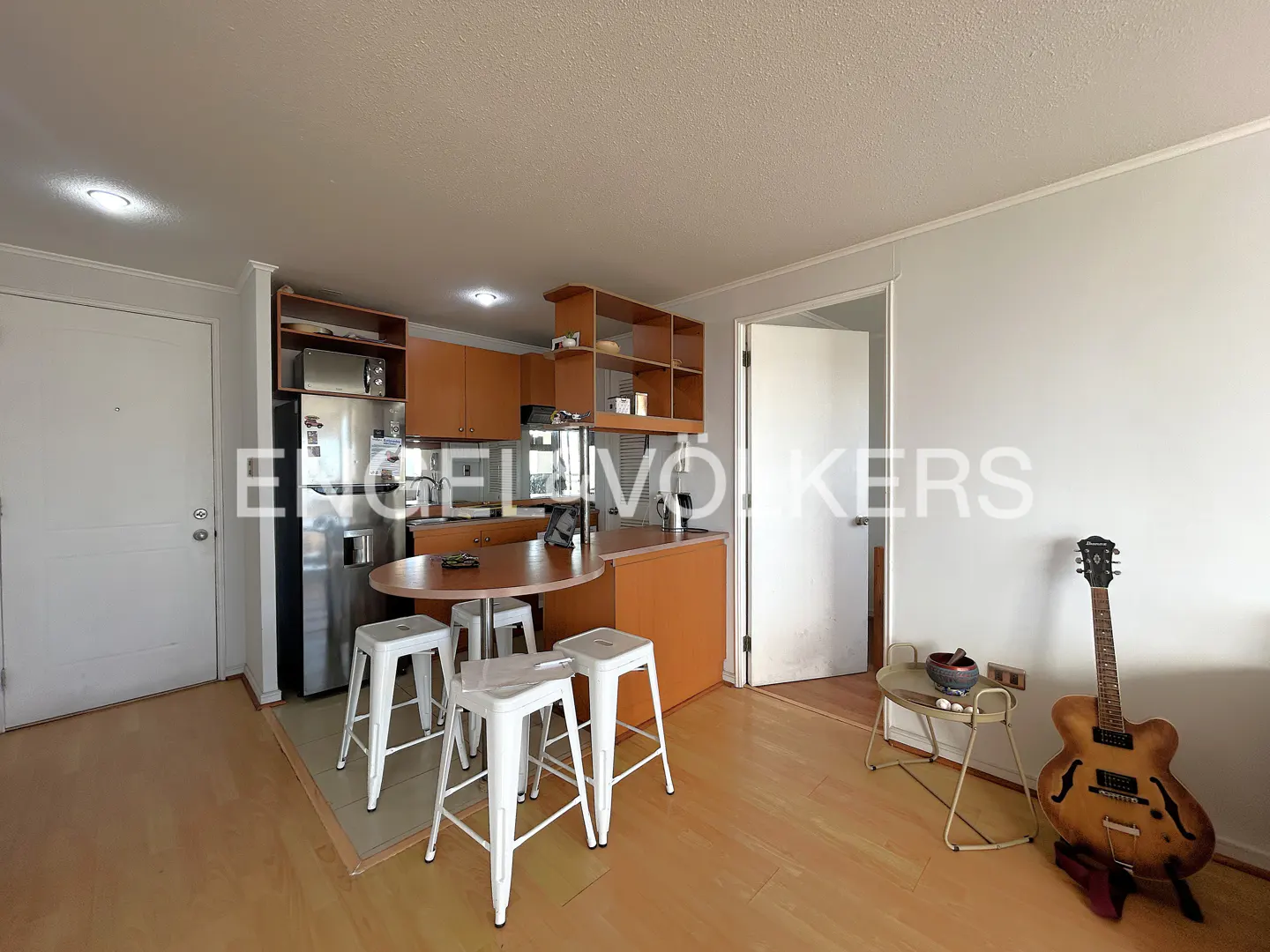 A bright, open-concept kitchen and living area with wood floors. Stainless steel appliances, white stools at a breakfast bar, and a guitar in the corner.