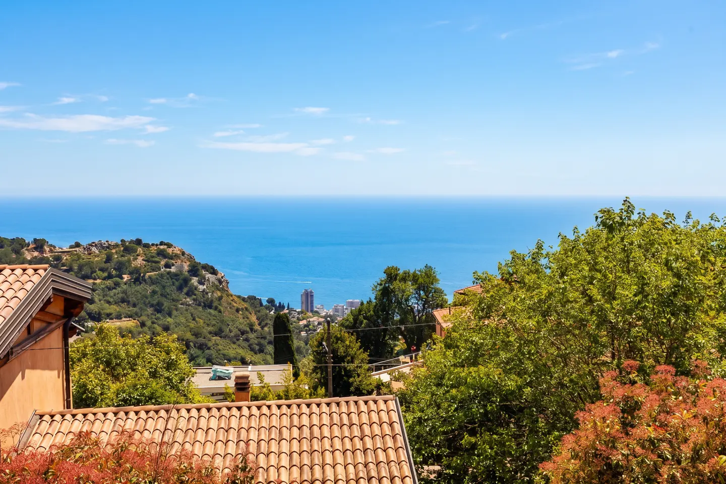 View of the Mediterranean Sea from a hillside with trees and terracotta rooftops under a blue sky.
