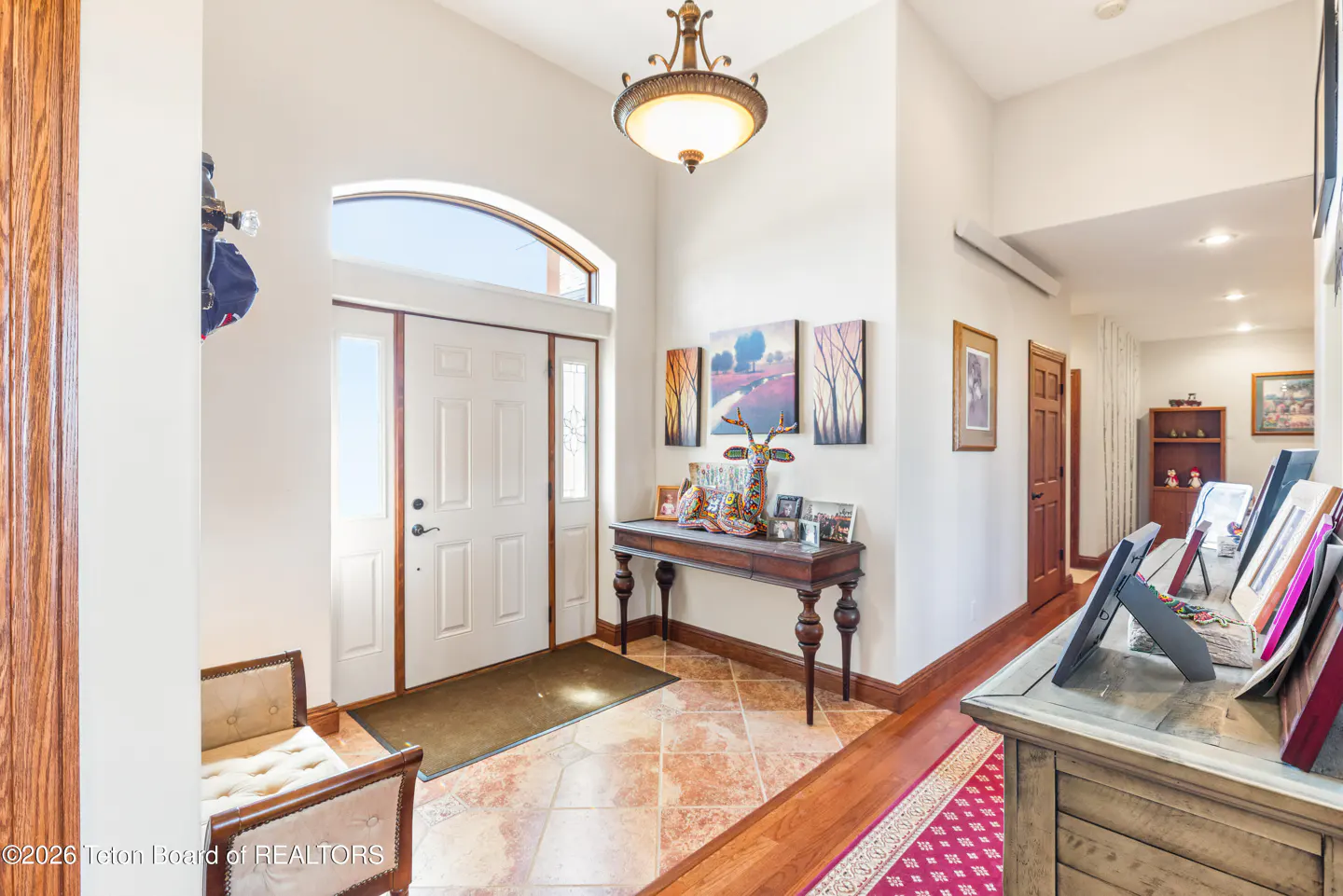 Bright foyer with white door, arched window, and bronze light fixture. A wooden table displays art and photos. Hallway extends into the home.