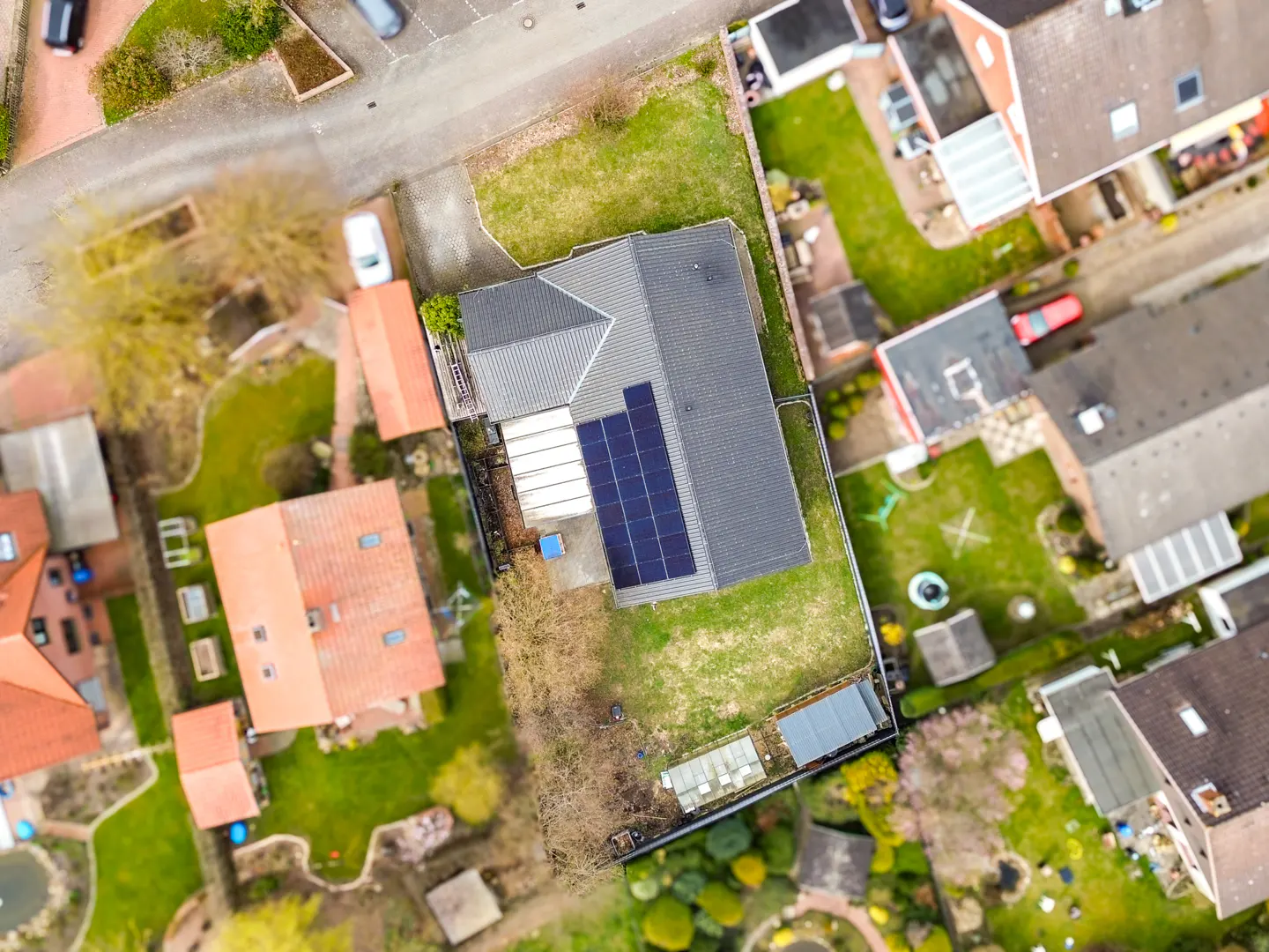 Aerial view of a gray roofed house with solar panels, surrounded by green lawns and other houses.