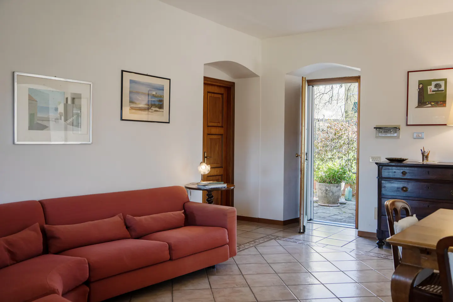 Living room with terracotta tile floor, red sofa, and open door to a garden patio. Artwork hangs on the white walls.