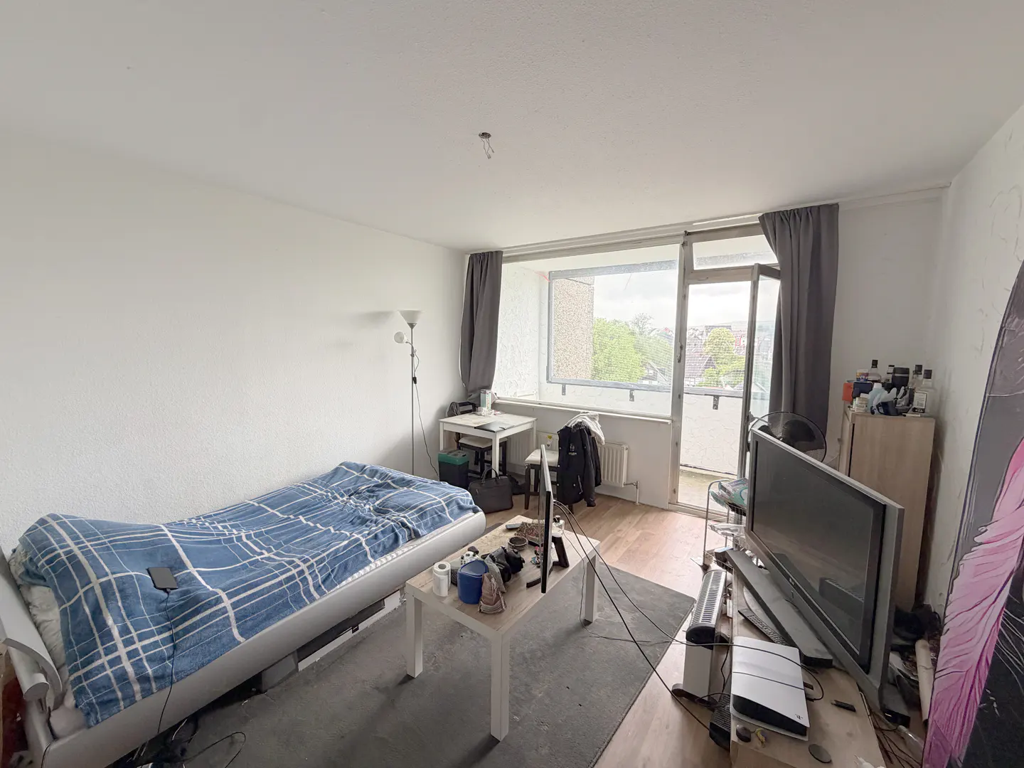 A wide shot of a cluttered studio apartment with a bed, TV, desk, and balcony. The walls are white, and the floor is wood.