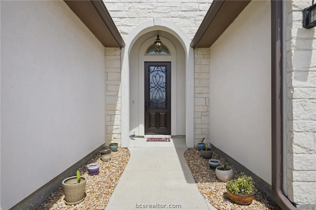 Front entrance of a home with a brown door, stone archway, and potted plants along a concrete walkway.