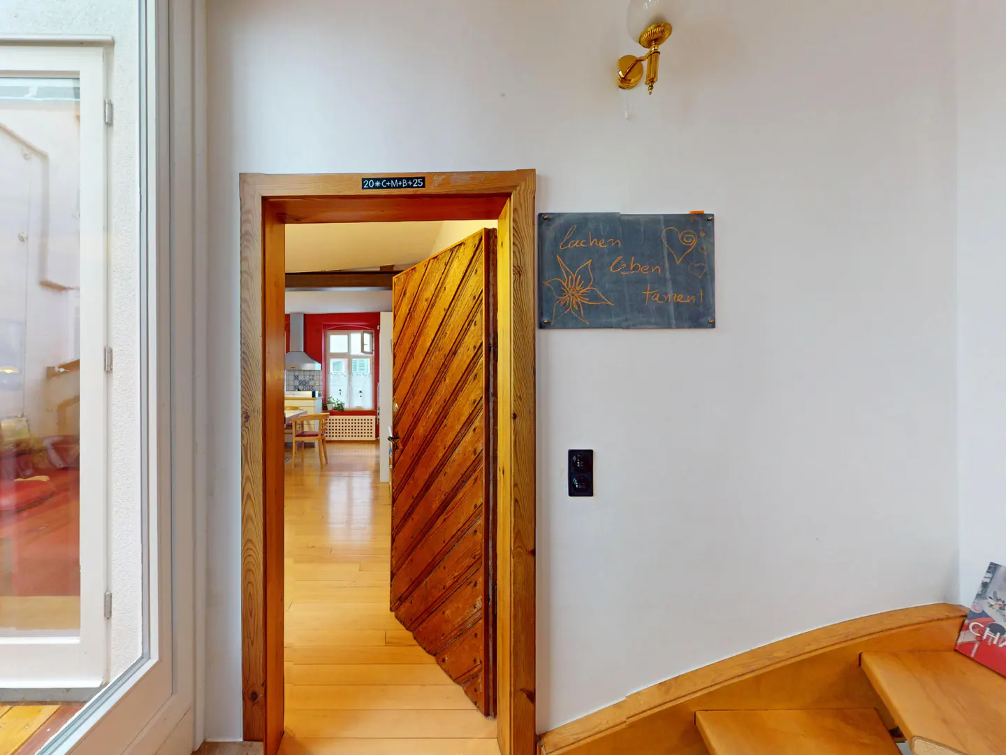 Interior view of a home with an open wooden door leading to a kitchen, a slate board with German text, and wooden stairs.