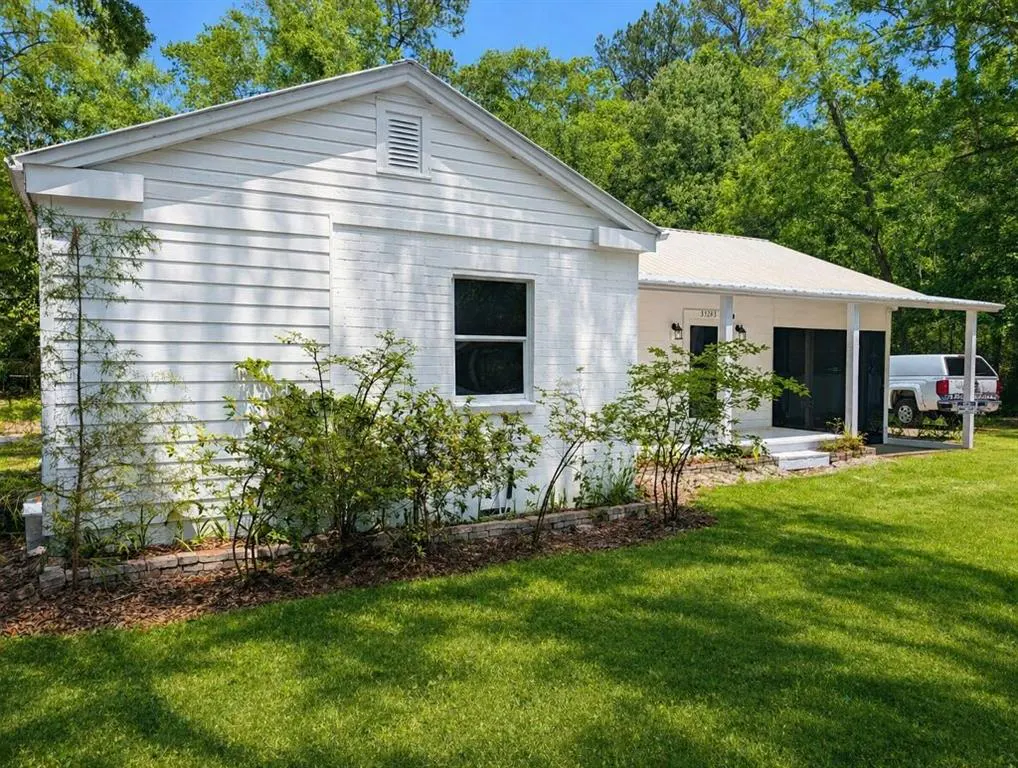Exterior view of a one-story white house with a metal roof, green lawn, and trees in the background. A white truck is parked under the house's porch.