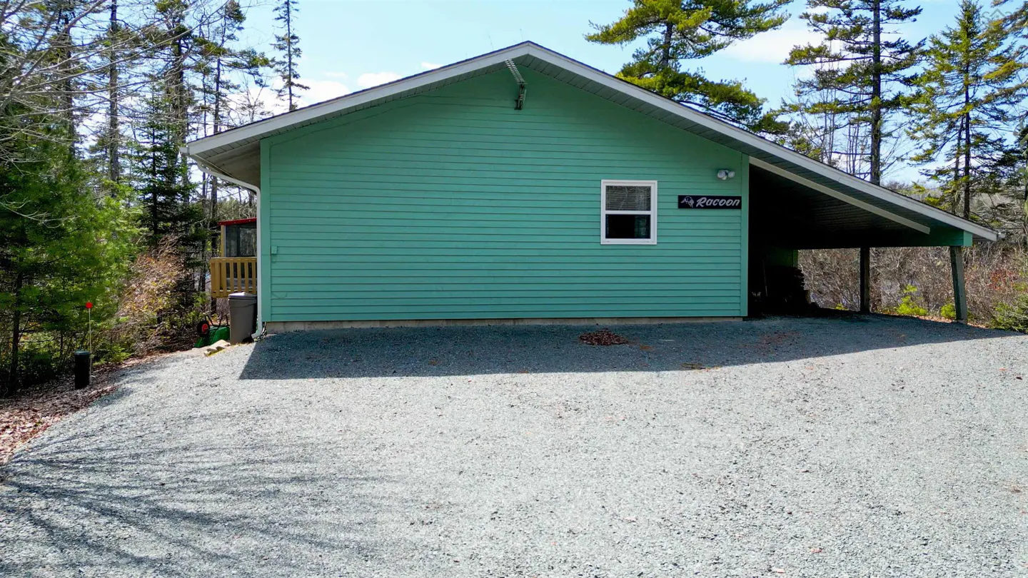 Exterior view of a light green house with a carport and a gravel driveway surrounded by trees. A sign reads "Racoon".