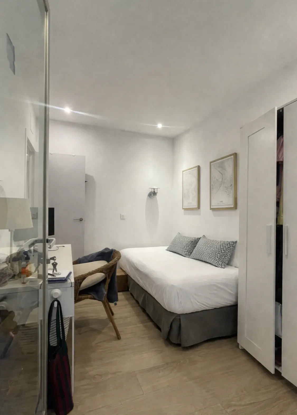 Bright bedroom with white walls, a bed with gray accents, and a desk with a wicker chair. Two framed pictures hang above the bed.