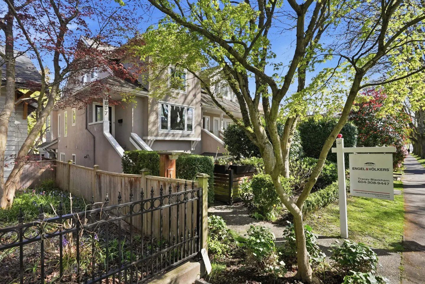 Exterior view of a two-story, taupe townhouse with a gray roof, surrounded by greenery and a wooden fence. An Engel & Völkers sign is on the right.