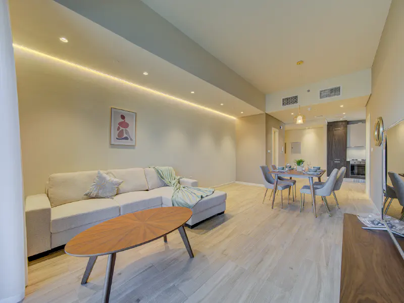 A bright, modern apartment interior with a beige sofa, wood coffee table, dining set, and kitchen in the background.