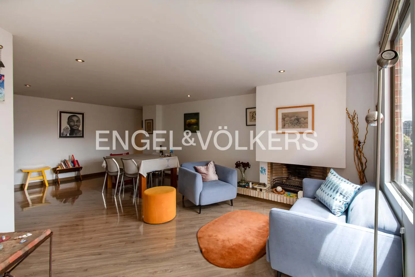 Open-concept living and dining area with light wood floors, blue sofas, and a white fireplace. Engel & Völkers logo overlay.