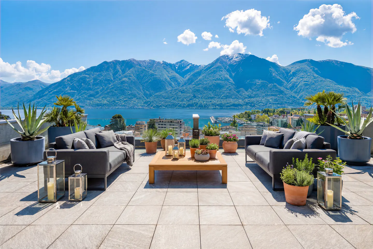 Rooftop patio with gray sofas, wood table, lanterns, and potted plants. Lake and mountains in the background under a blue sky.