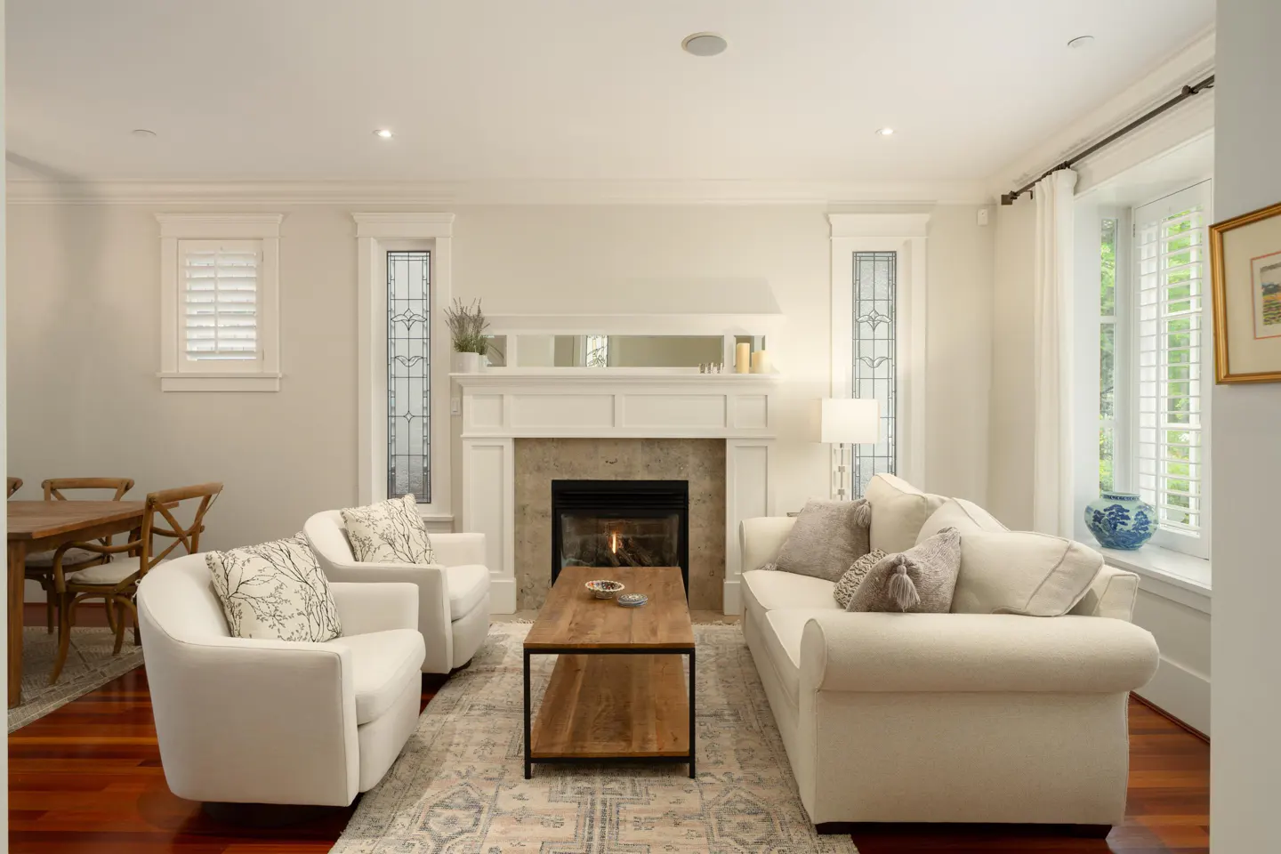 Living room with white sofa and chairs, fireplace, wood coffee table, and patterned rug. Dining table visible in background.