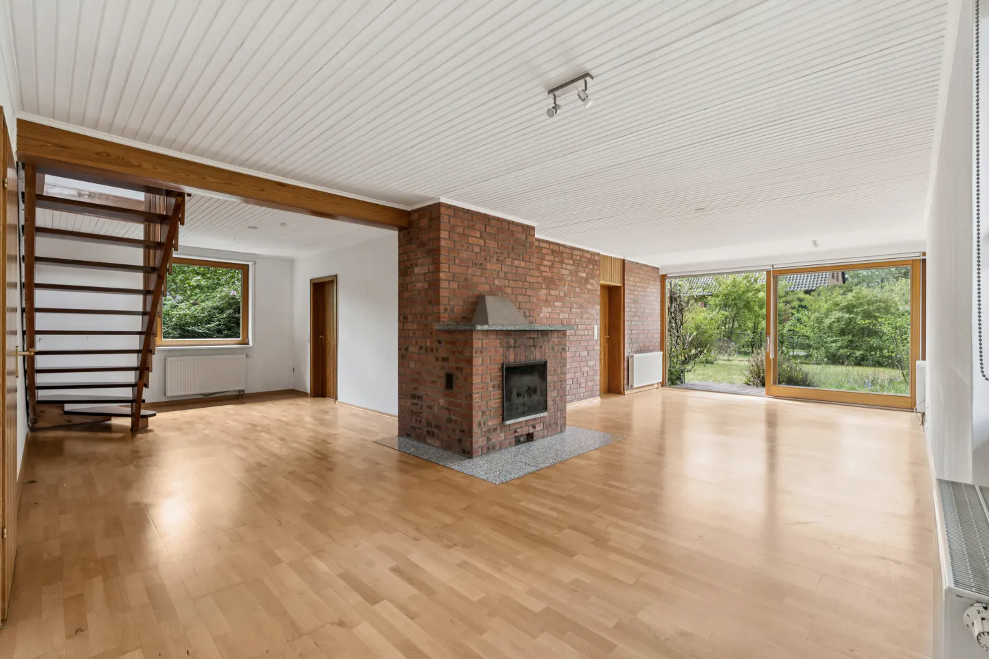 Bright, empty living room with wood floors, a brick fireplace, and a staircase. Large sliding glass doors lead to a green yard.