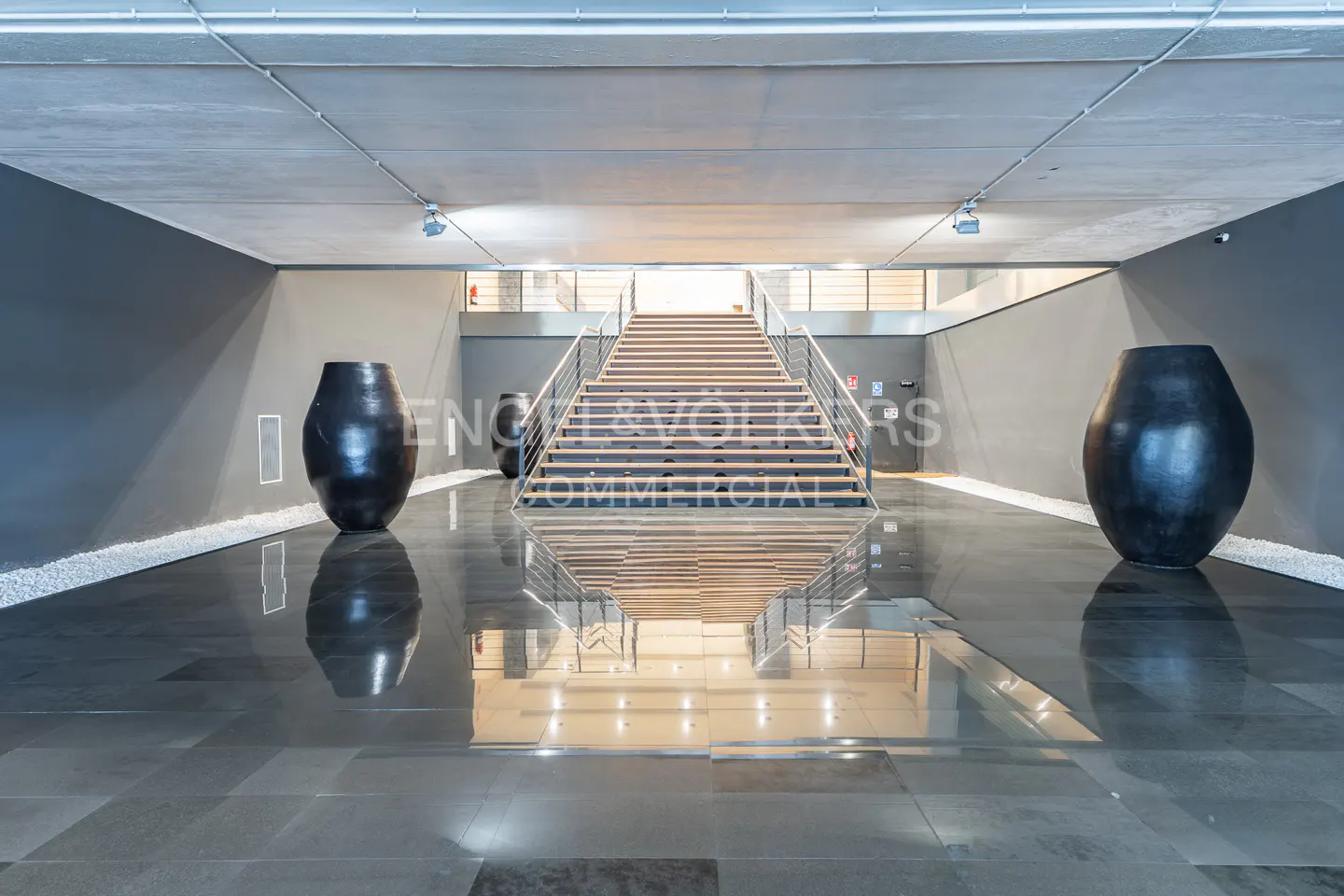 Interior view of a modern lobby with gray walls, a staircase, and two large black vases. The floor is shiny and reflects the light.