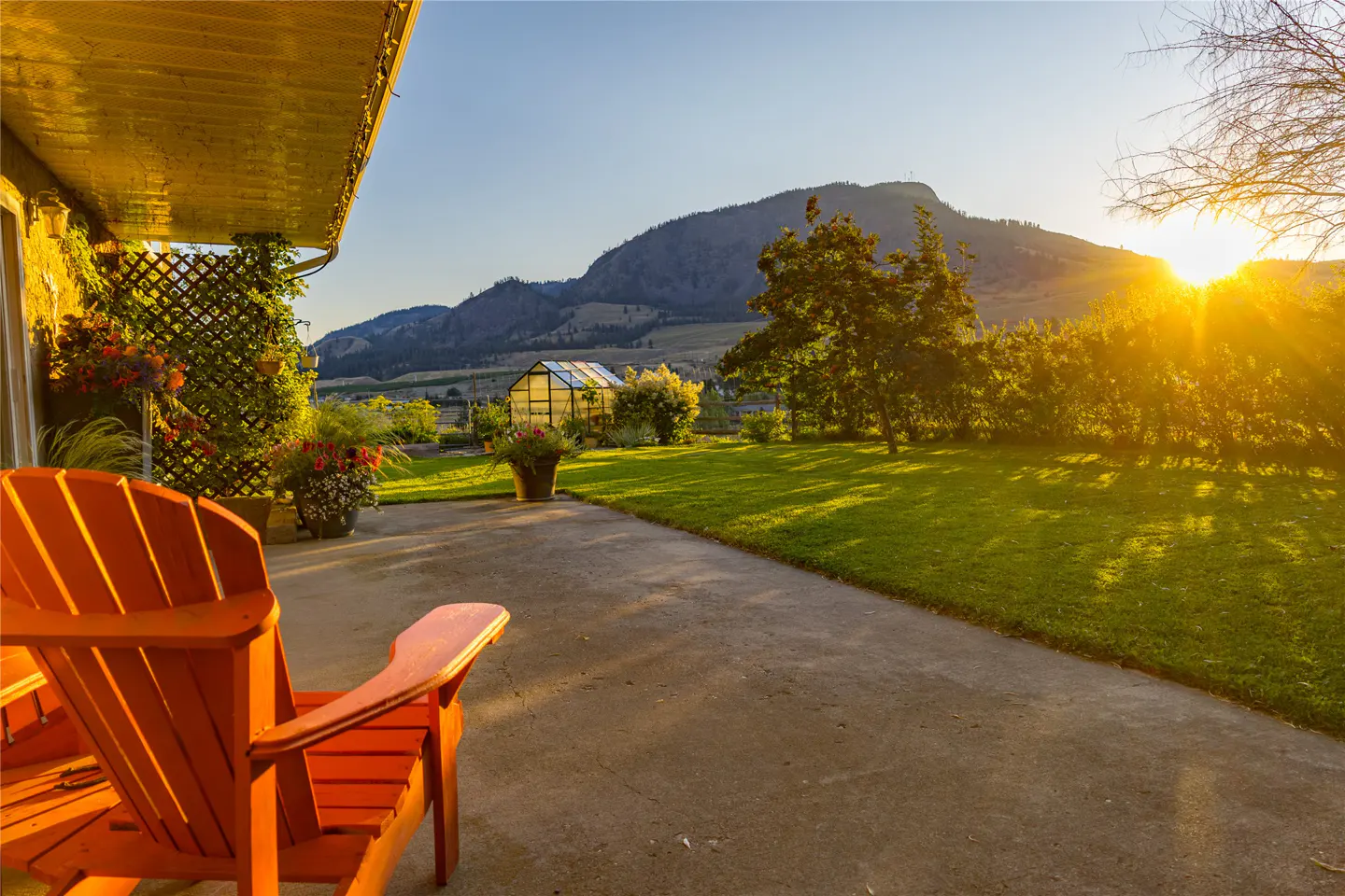Orange Adirondack chairs sit on a patio overlooking a green lawn, greenhouse, and mountain at sunset. Flowers decorate the porch.