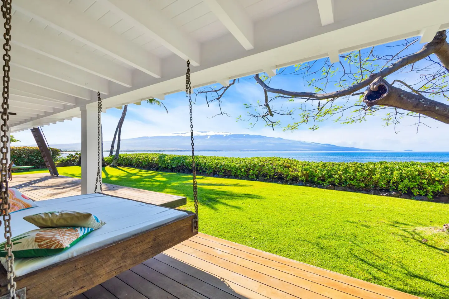 A hanging daybed on a wooden deck overlooks a lush green lawn, the ocean, and a distant mountain under a blue sky.