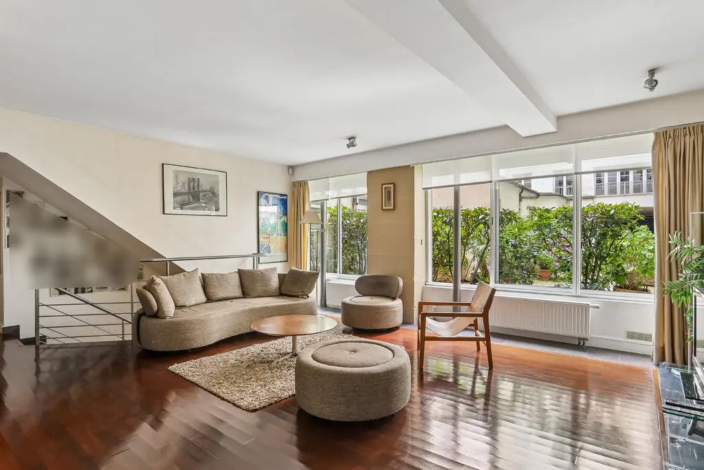 Bright living room with dark hardwood floors, beige sofa, round coffee table, ottoman, and large windows overlooking greenery.