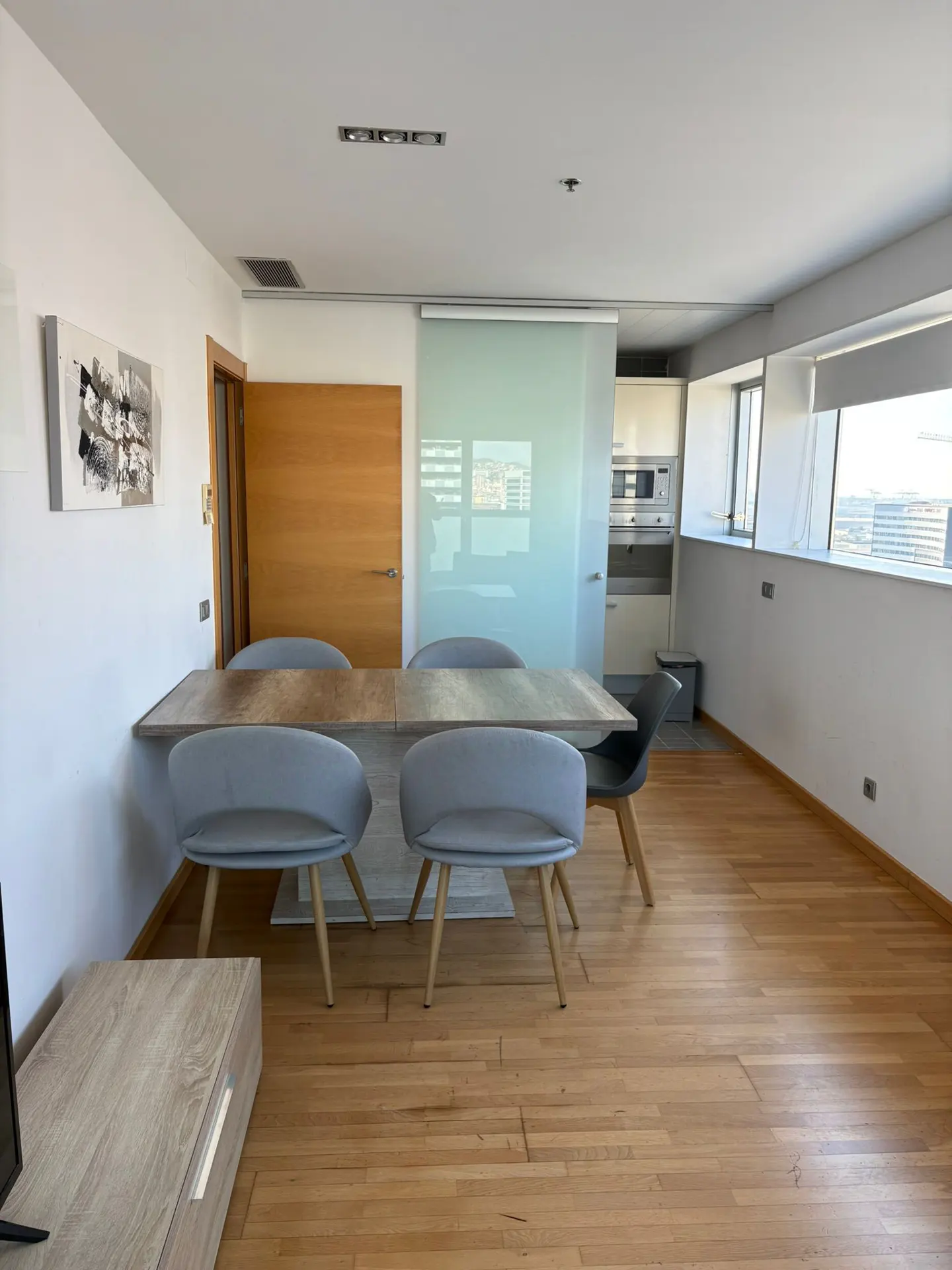 A bright, modern dining area with a wooden table, gray chairs, and hardwood floors. A sliding glass door leads to the kitchen.