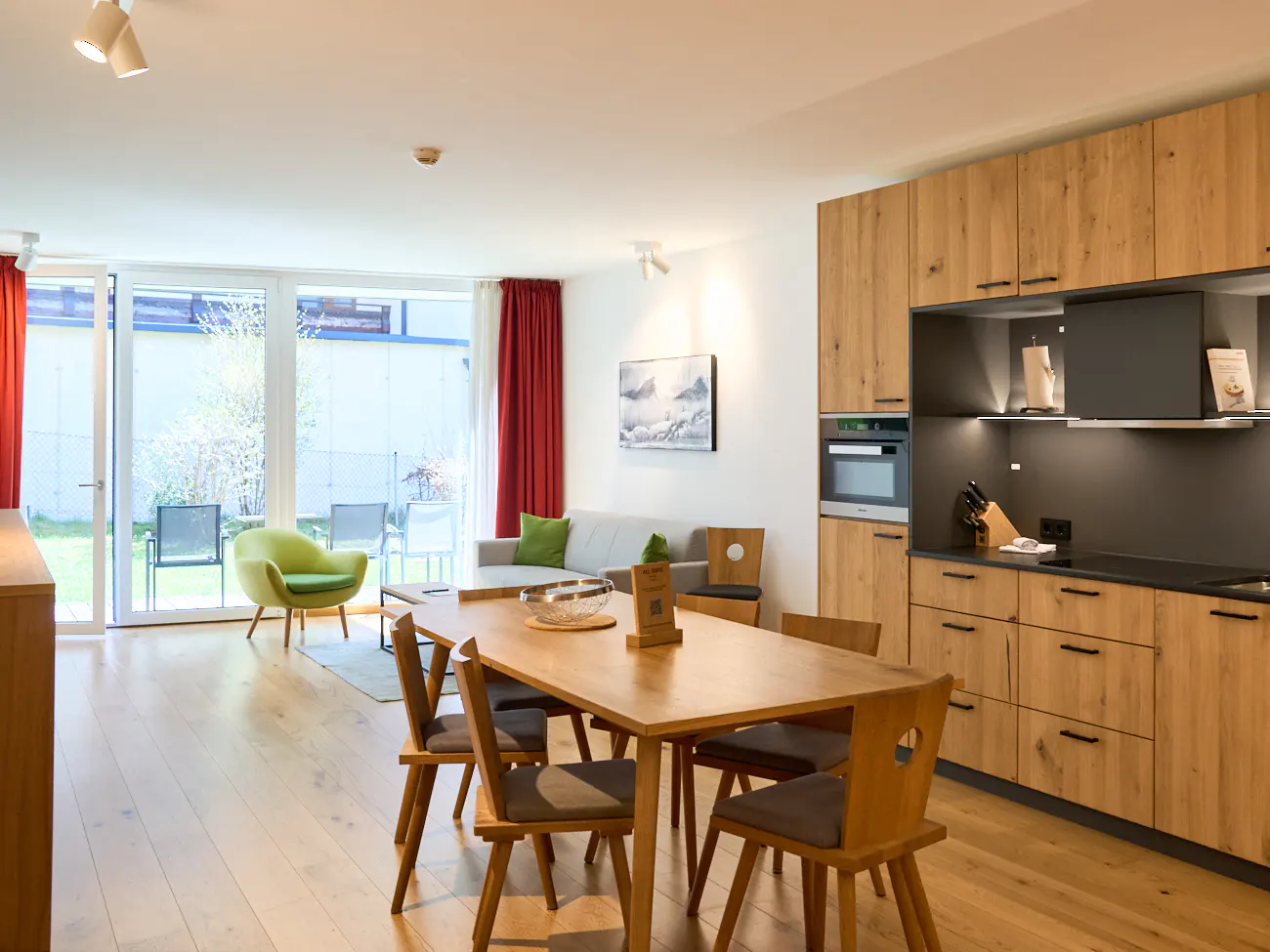 Bright apartment interior with wood floors, a dining table with six chairs, and a modern kitchen with wood cabinets. Sliding glass doors lead to a patio.