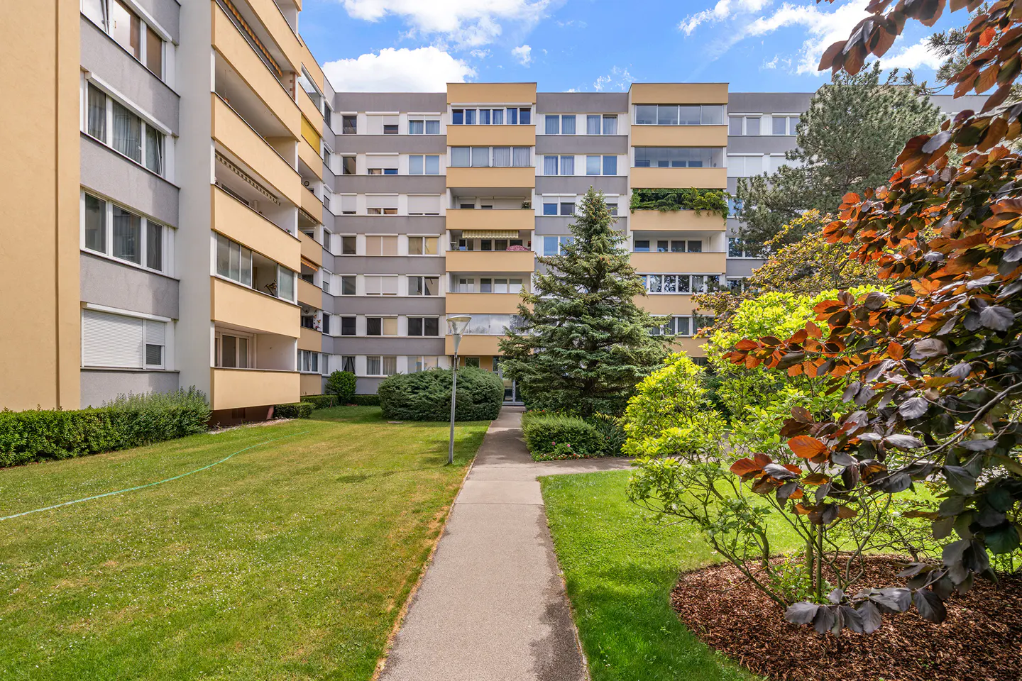 Exterior view of a modern apartment building with a green lawn and trees. The building is gray and yellow with balconies.