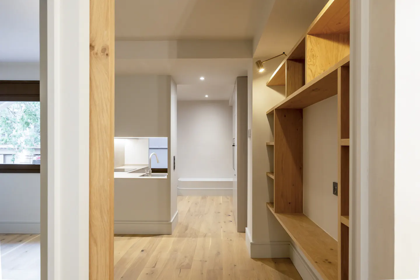 Interior view of a modern home with light wood floors, white walls, and a built-in wooden shelving unit. A glimpse of the kitchen is visible.