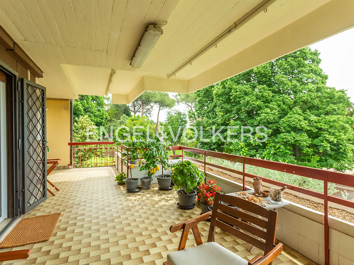 A covered balcony with a red railing overlooks lush green trees. Potted plants and a table with snacks add to the inviting outdoor space.