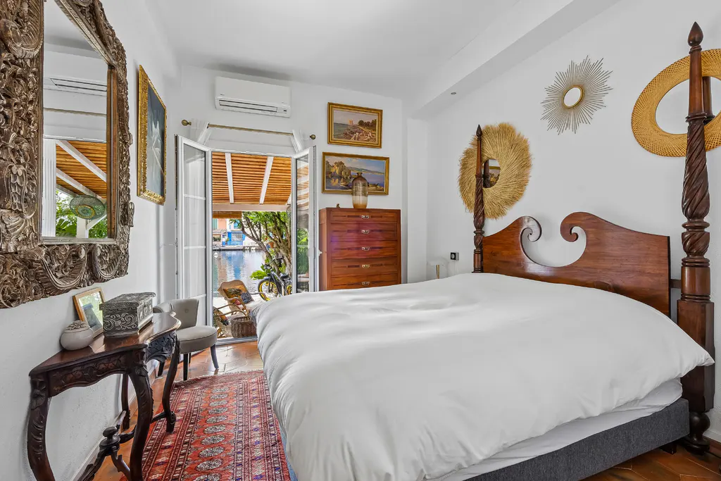 Bedroom with white walls, a four-poster bed, and an open door leading to a canal view. Ornate mirror and table on the left.