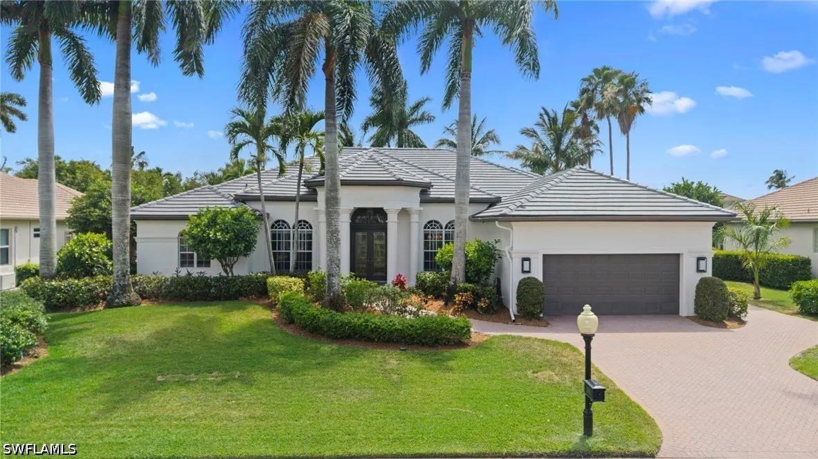 A white single-story house with a gray roof, palm trees, green lawn, and a brick driveway.