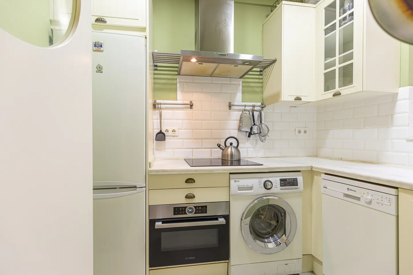 A compact kitchen with white cabinets, appliances, and subway tile. A stainless steel kettle sits on the stovetop.