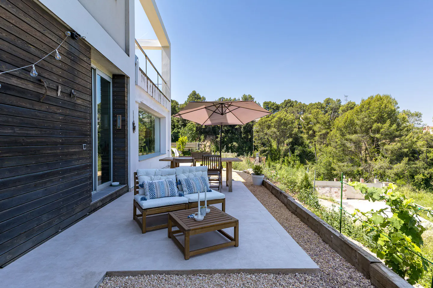 Outdoor patio with a wooden sofa, table, and umbrella. Lush green trees in the background.
