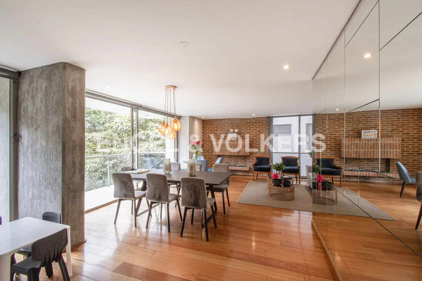 Open-concept living and dining area with wood floors, brick wall, and large windows. Dining table with gray chairs. Modern decor.
