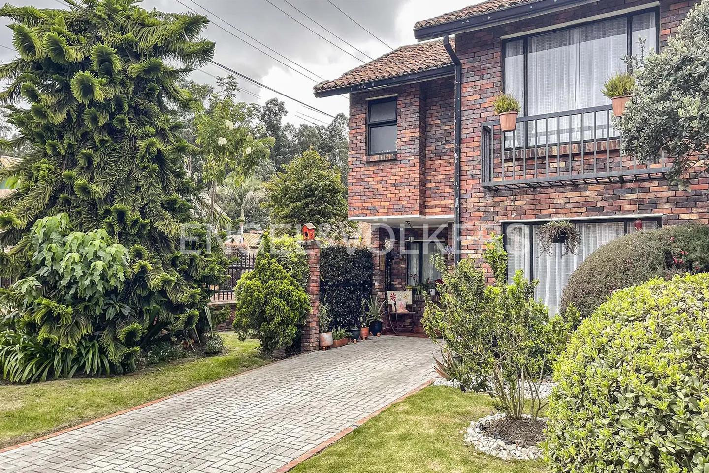 A two-story brick house with a tiled roof, a balcony, and a paved driveway surrounded by lush greenery.
