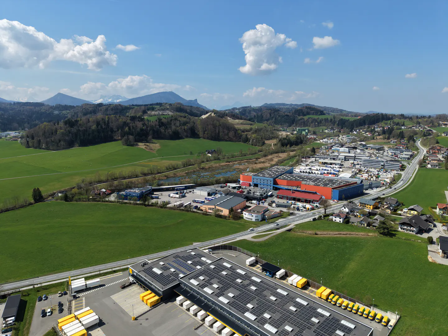 Aerial view of a commercial property with solar panels, loading docks, and yellow trucks, set against a backdrop of green fields, mountains, and a blue sky.