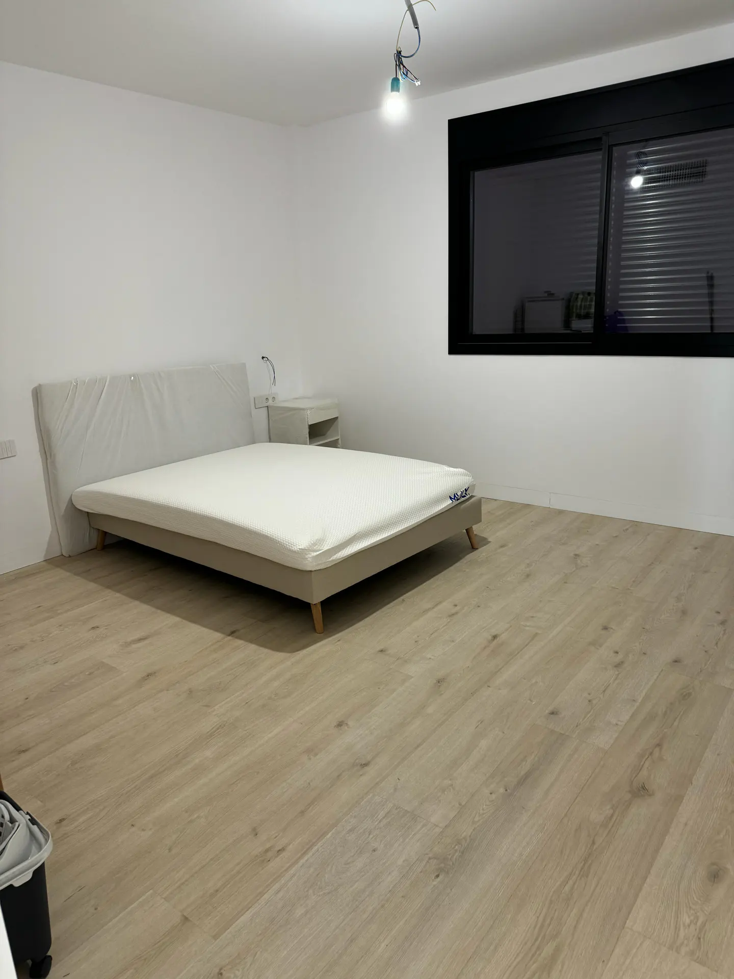 A bedroom with white walls, a light wood floor, and a bed with a white mattress. A black-framed window is on the right wall.