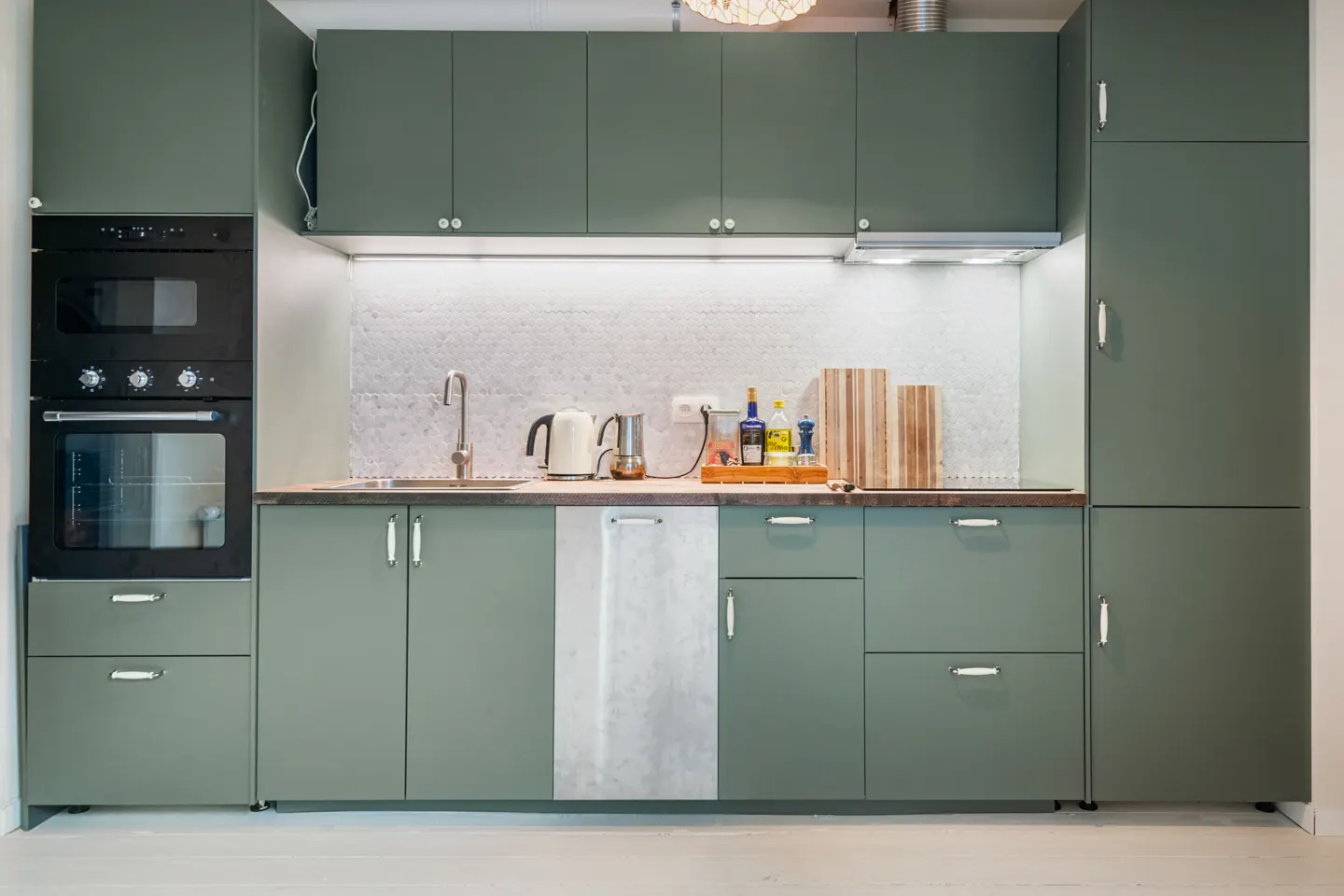 A modern kitchen with sage green cabinets, a black oven, and a white backsplash. A kettle and cutting boards sit on the counter.