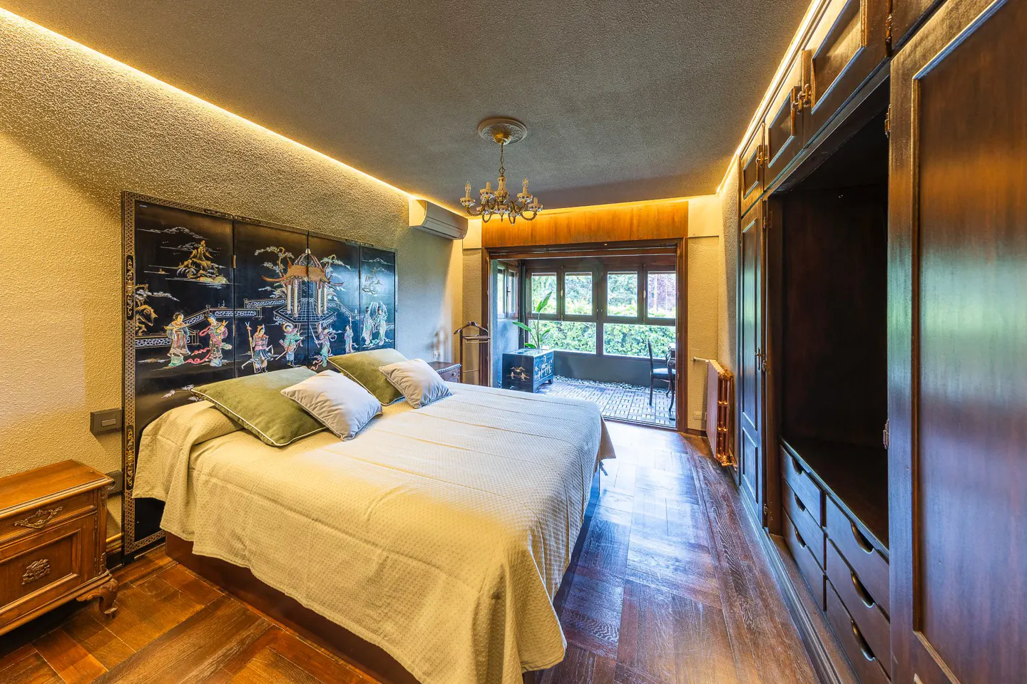 Bedroom with a large bed, black headboard with Asian art, and a balcony with a view of greenery. A dark wood wardrobe stands to the side.