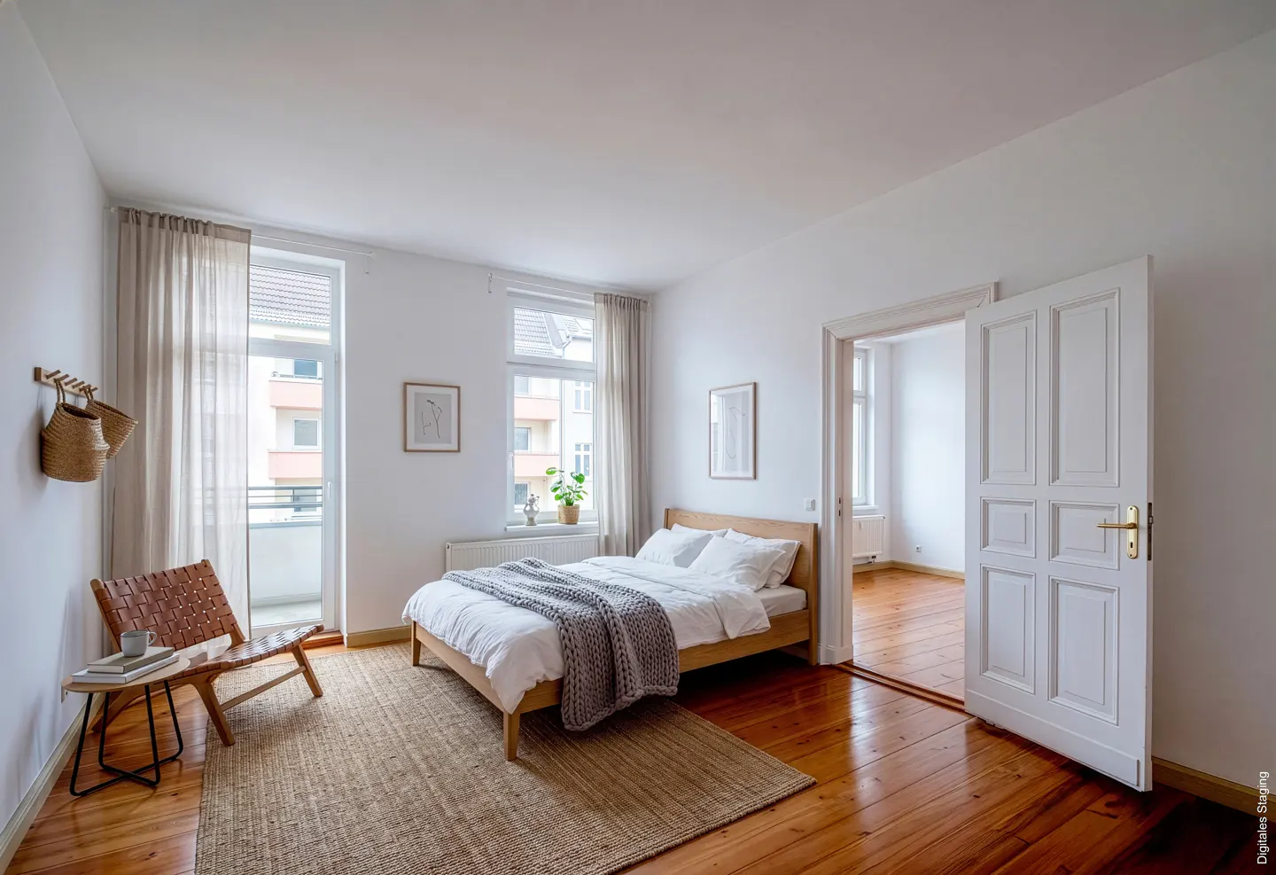 Bright bedroom with wood floors, white walls, and a jute rug. A wooden bed is covered in white linens and a gray throw. A leather chair sits near a balcony.