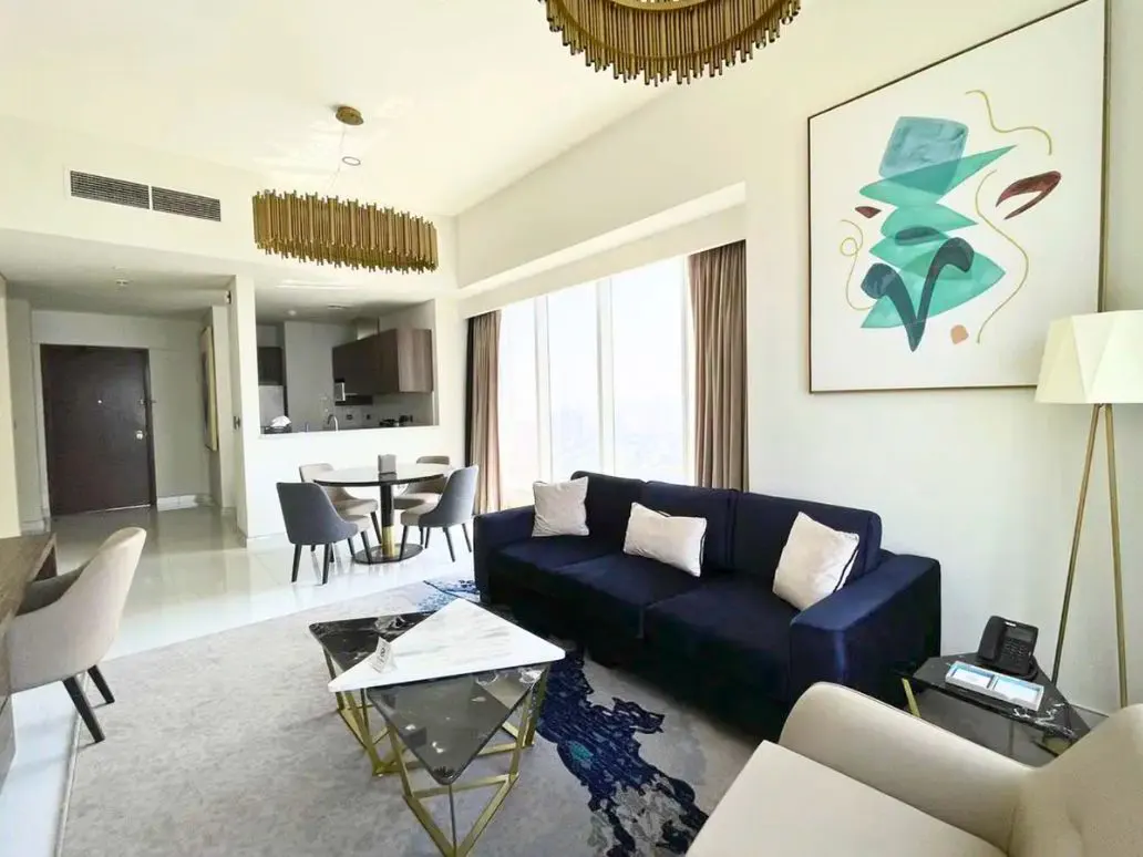 A modern living room with a blue sofa, abstract art, and a gold chandelier. A dining table and kitchen are visible in the background.