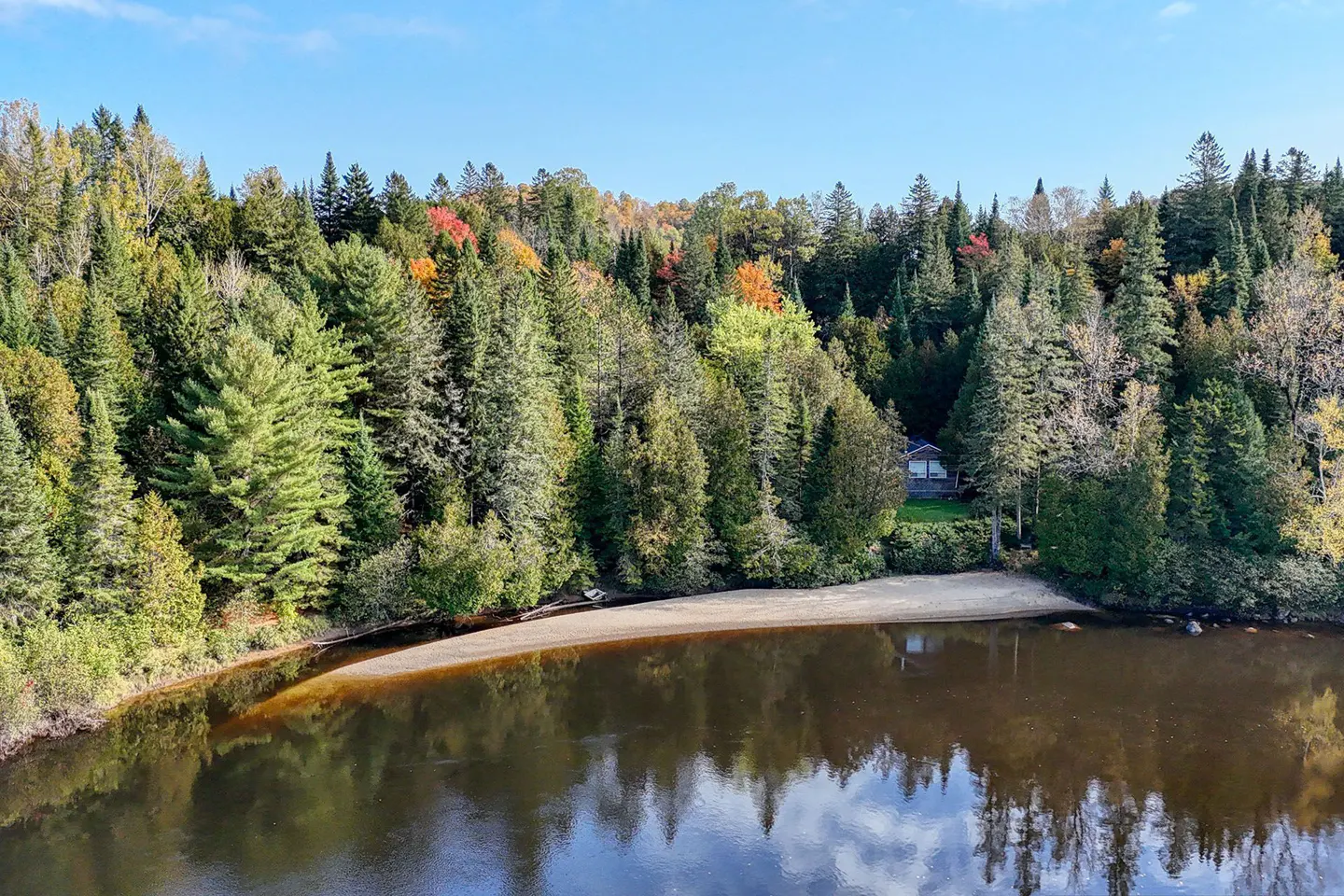 A secluded cabin nestled in a dense, colorful autumn forest beside a calm river with a sandy shoreline.