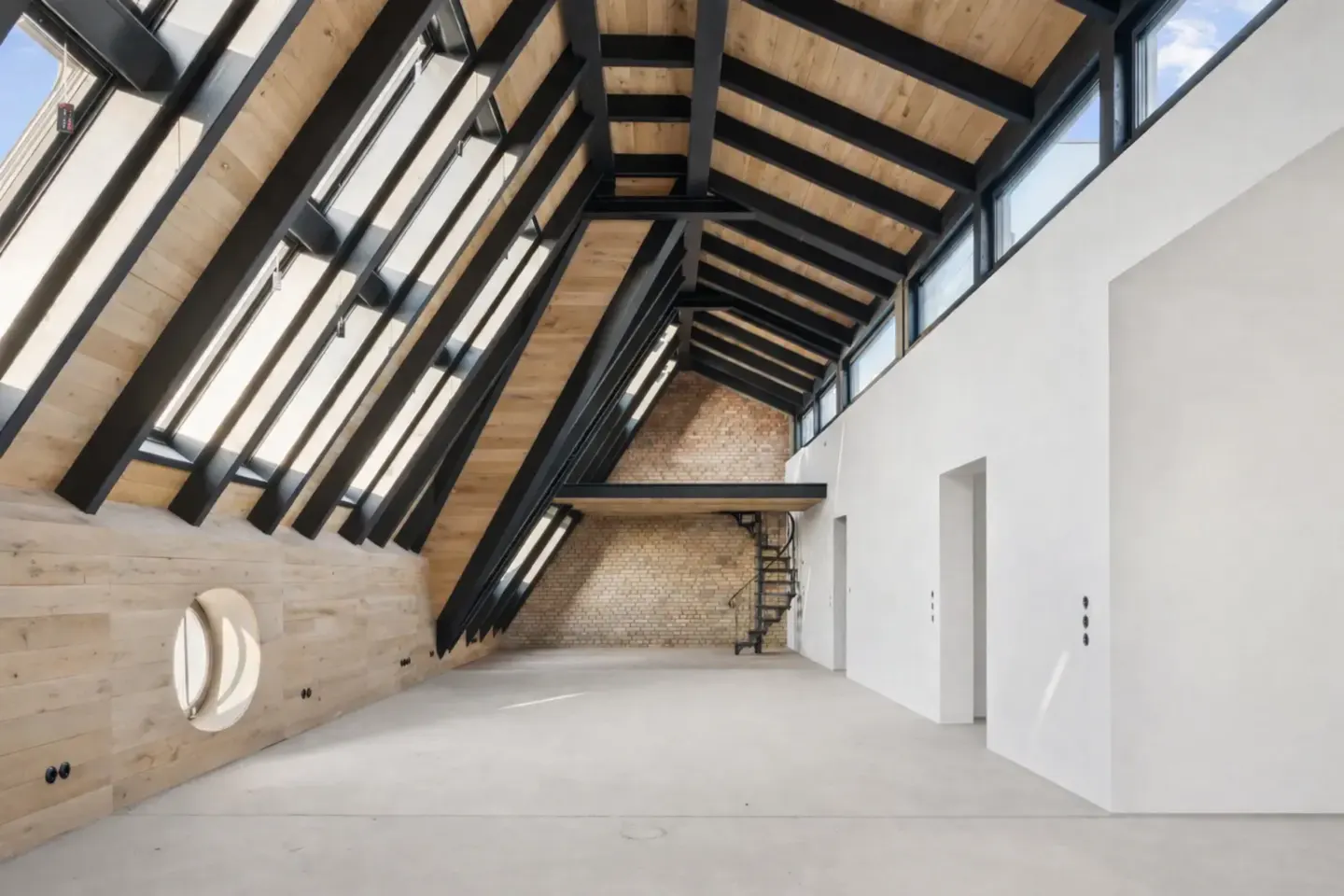 An interior view of a modern loft with a high, angled ceiling, black beams, and light wood panels. A spiral staircase leads to a small platform. White walls and concrete floors.