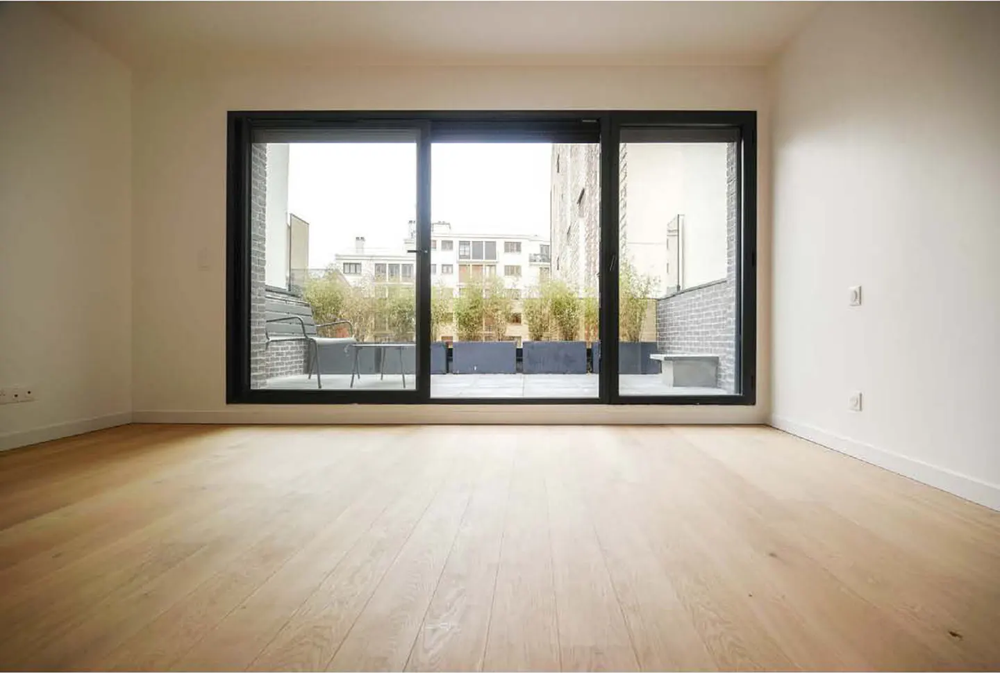 Empty room with light wood floors and large black-framed sliding glass doors opening to a balcony with plants.