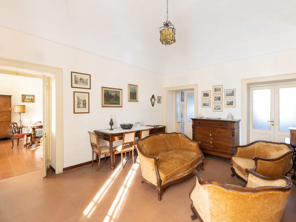 A living room with terracotta floors, white walls, and antique furniture. A dining table, dresser, and gold velvet sofas are visible.