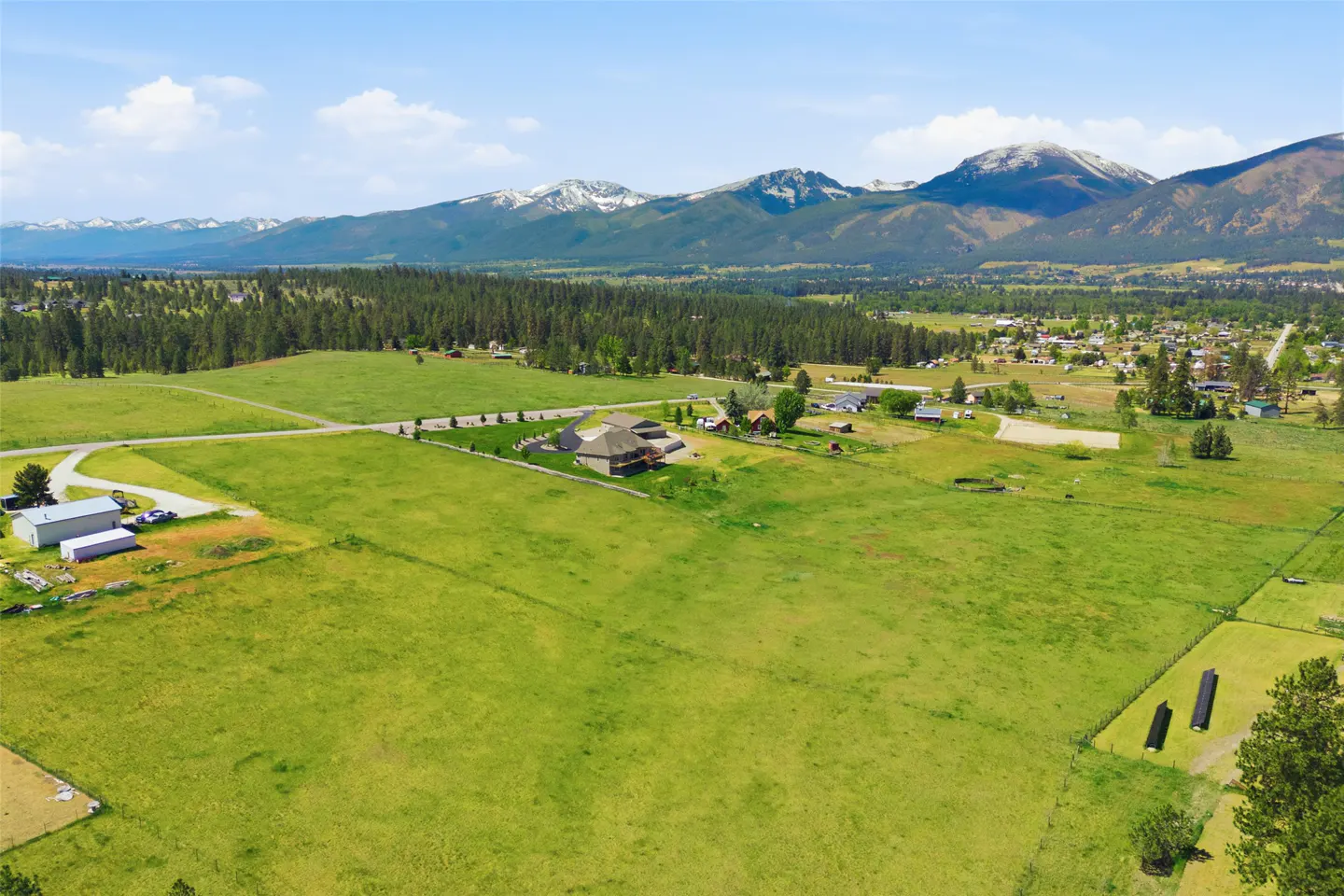 Aerial view of a large green field with a house, trees, and snow-capped mountains in the background under a blue sky.